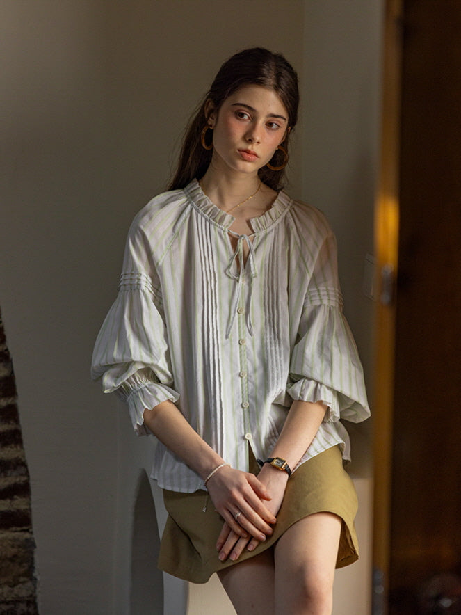 A young woman seated indoors, wearing a light green and white striped blouse with puffed sleeves and a ruffled neckline. Her relaxed pose and gentle expression evoke a calm, cottagecore-inspired atmosphere.