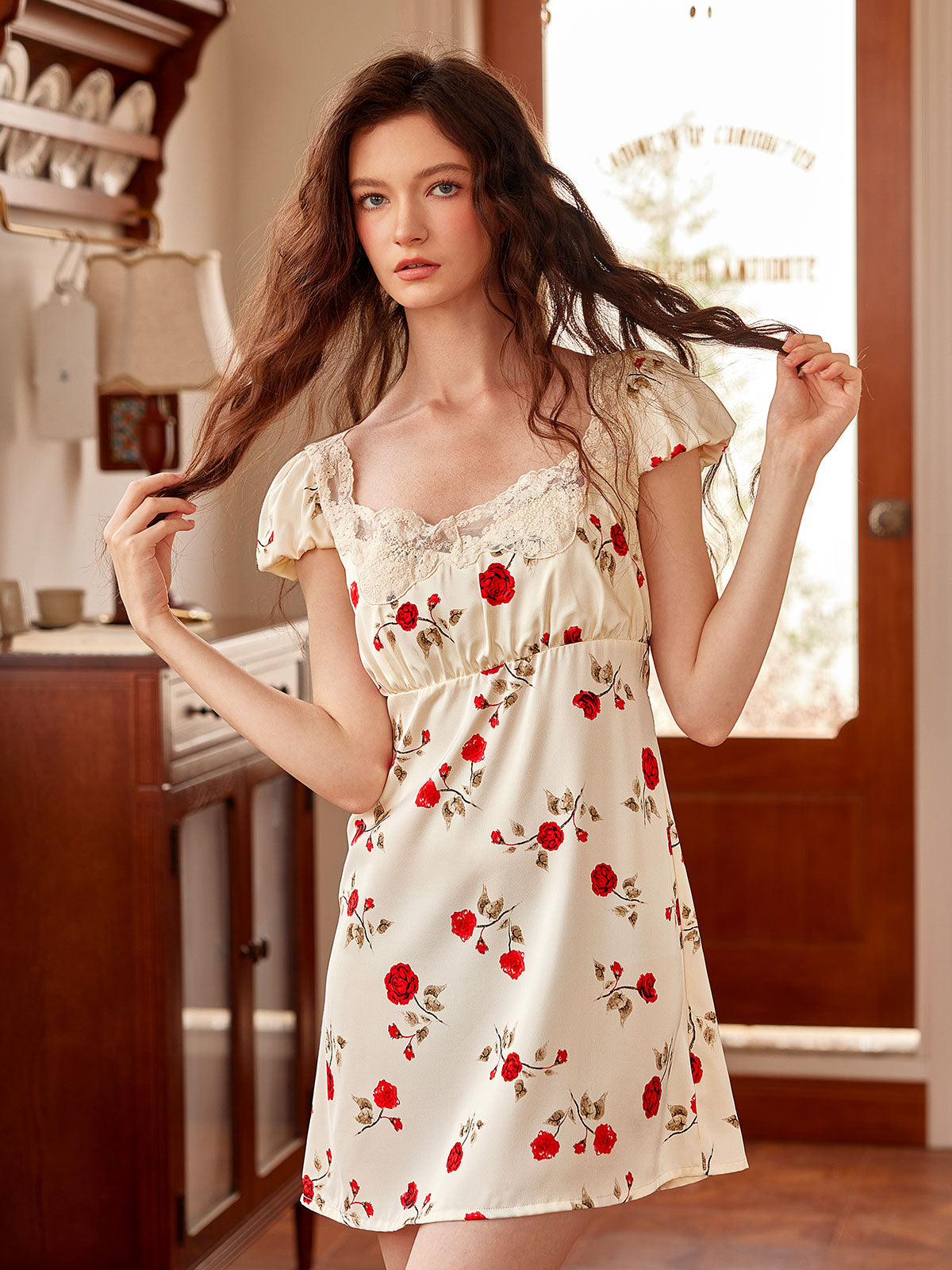 In a soft cottagecore mood, the model plays with her wavy hair while dressed in a cream nightgown adorned with red roses and puff sleeves.