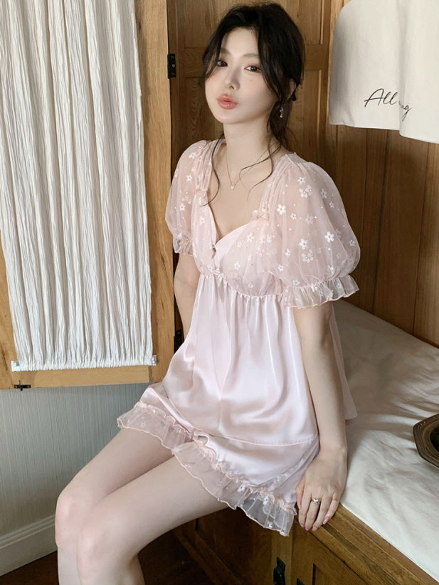 A woman in a pink coquette pajama set with daisy-embroidered mesh sleeves sits by a wooden cabinet, softly pouting in a romantic vintage mood.