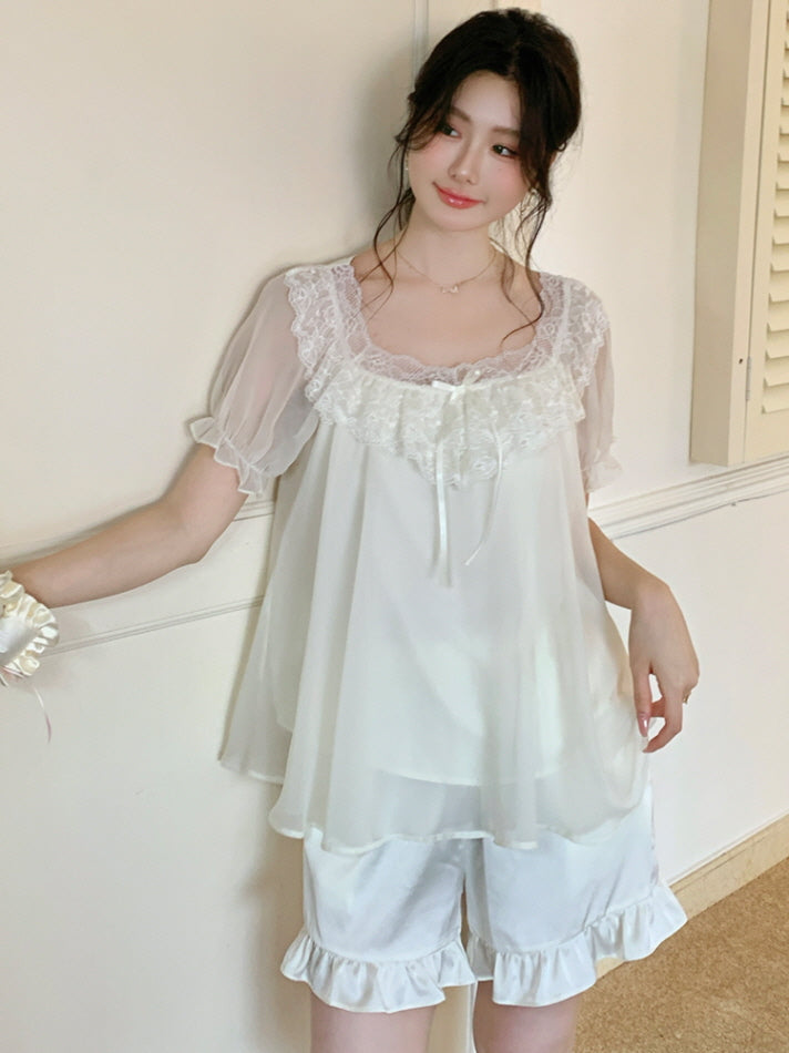 Woman leaning on a bedframe while wearing a layered ivory coquette fairy pajama set with lace neckline and puff sleeves.
